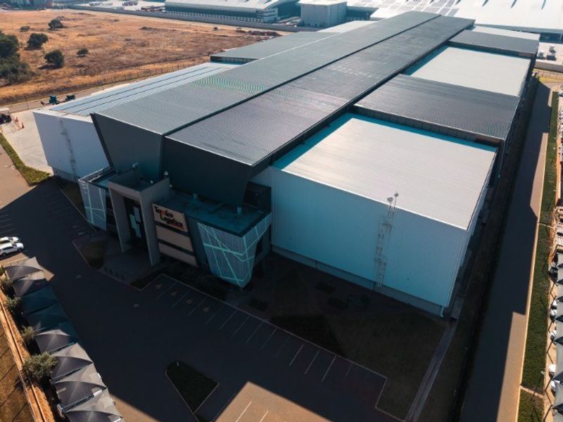 Aerial view of a large industrial warehouse facility featuring a modern metal roof, extensive parking area, and secure perimeter fencing.
