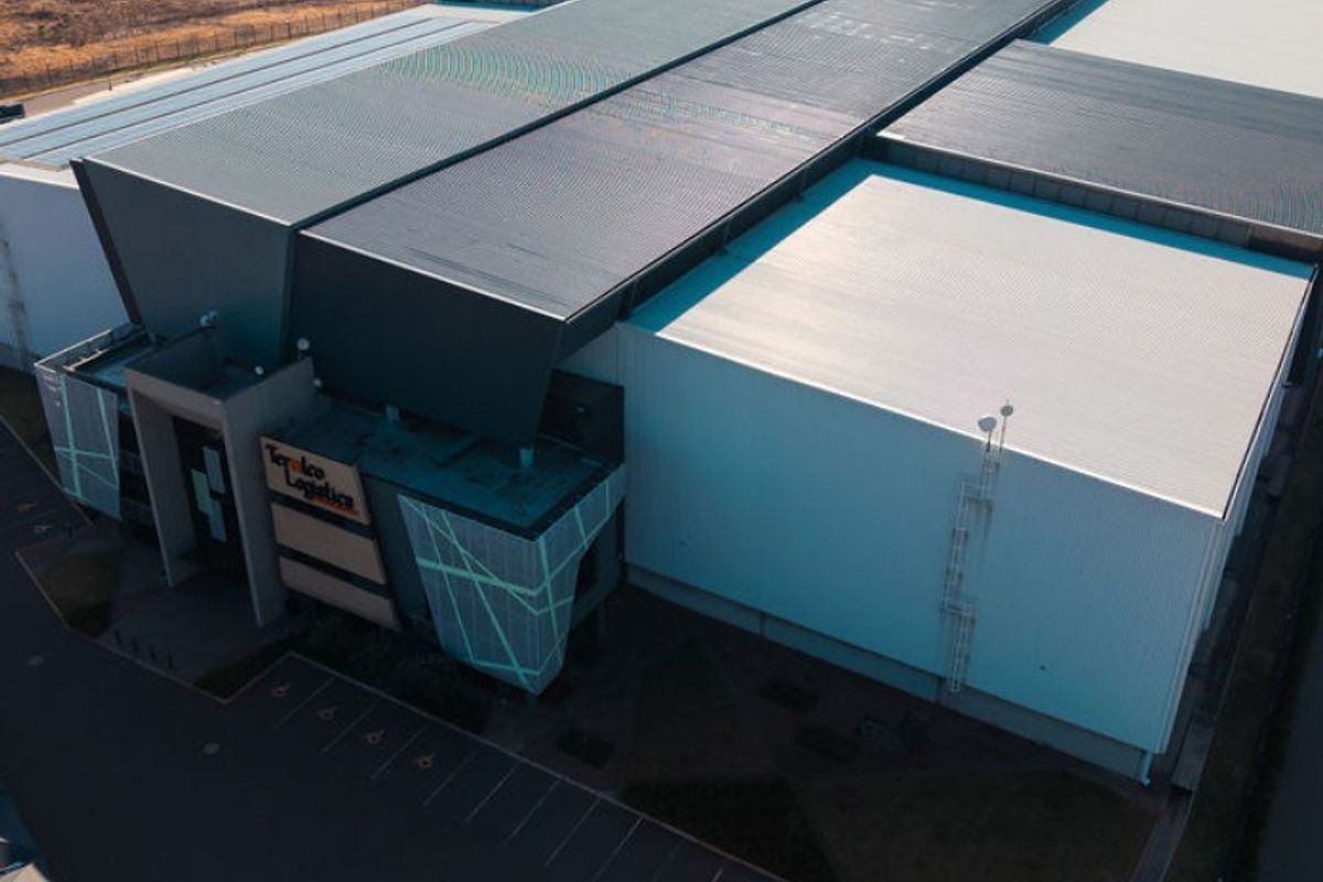 Aerial view of a modern industrial logistics facility featuring a large metal roof structure and dedicated asphalt parking area.
