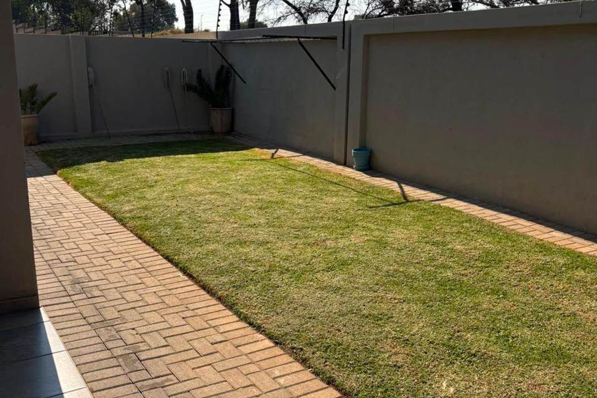 A well-maintained rear garden featuring a neat lawn, paved walkway, and secure perimeter wall with electric fencing.