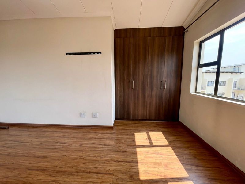 A bright, empty bedroom featuring wood-look laminate flooring, built-in dark wood cupboards, and a large window providing natural light.
