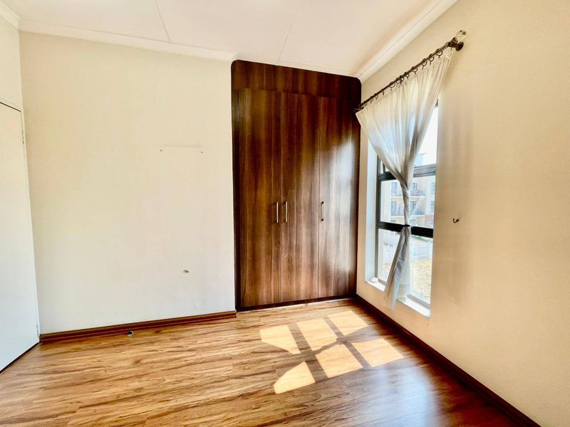 A bright, empty bedroom featuring warm-toned laminate flooring, a large built-in wooden cupboard, and a window allowing for natural light.