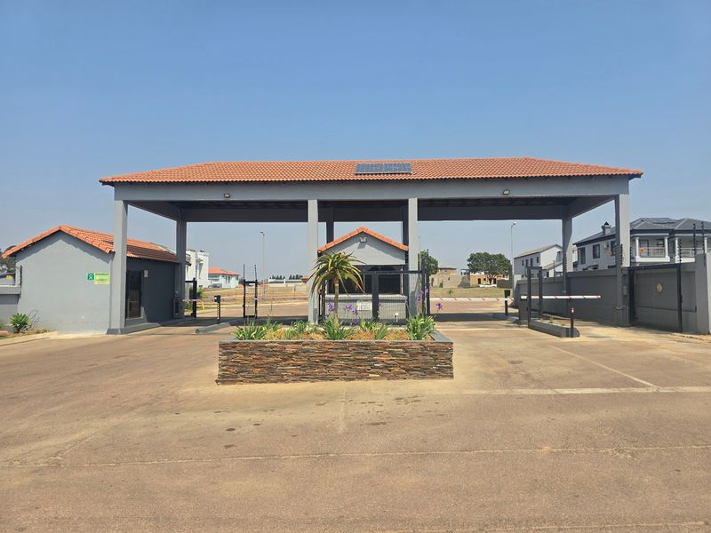 A well-maintained residential estate entrance featuring a security gatehouse, boom gates, and a stone-clad central planter under a covered roof structure.