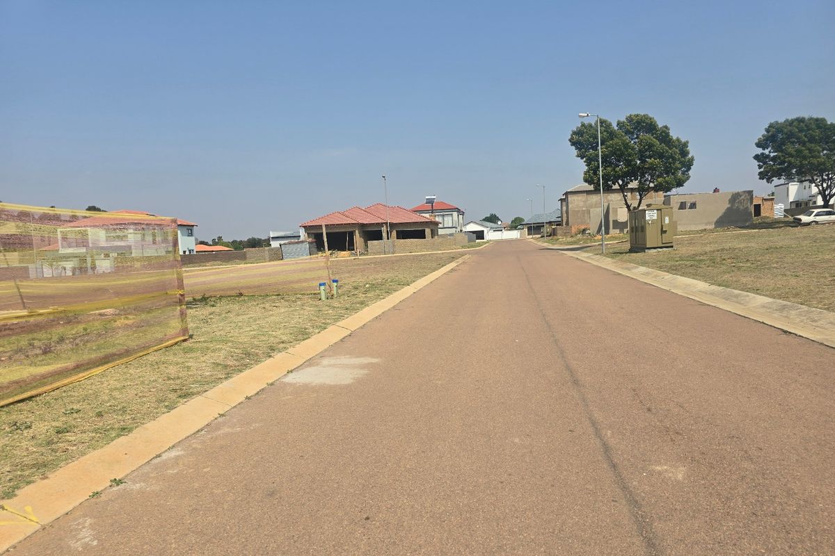 A view of a paved residential street in a developing neighborhood with surrounding vacant plots and nearby construction.