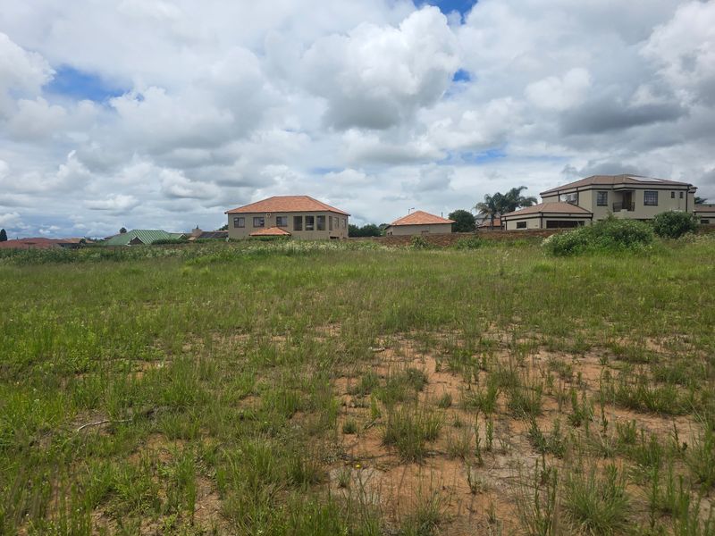 A view of a vacant residential plot of land with surrounding neighborhood houses and a perimeter wall in the background.