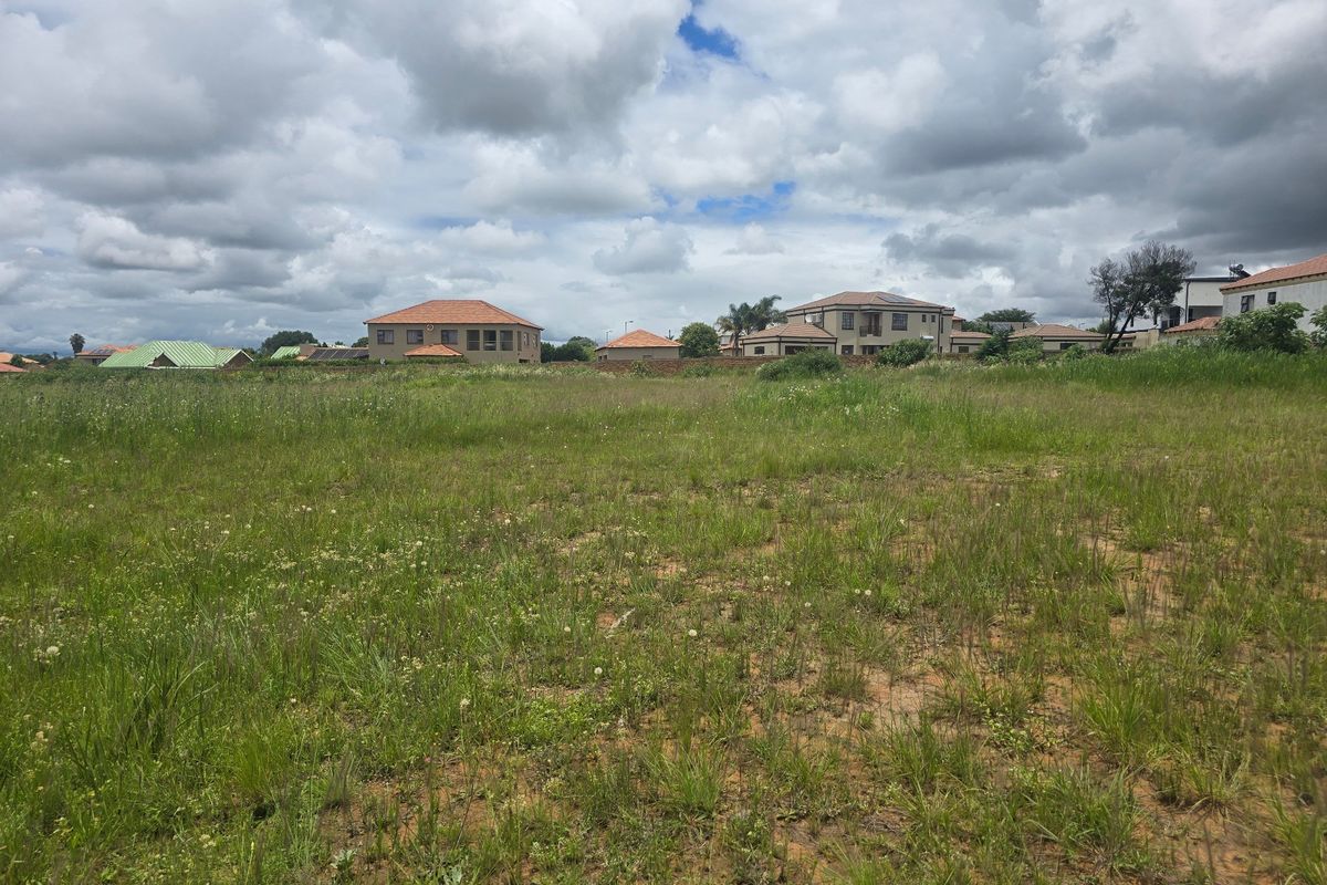 A vacant residential plot of land featuring natural grass coverage and a perimeter wall, situated within an established residential neighborhood.