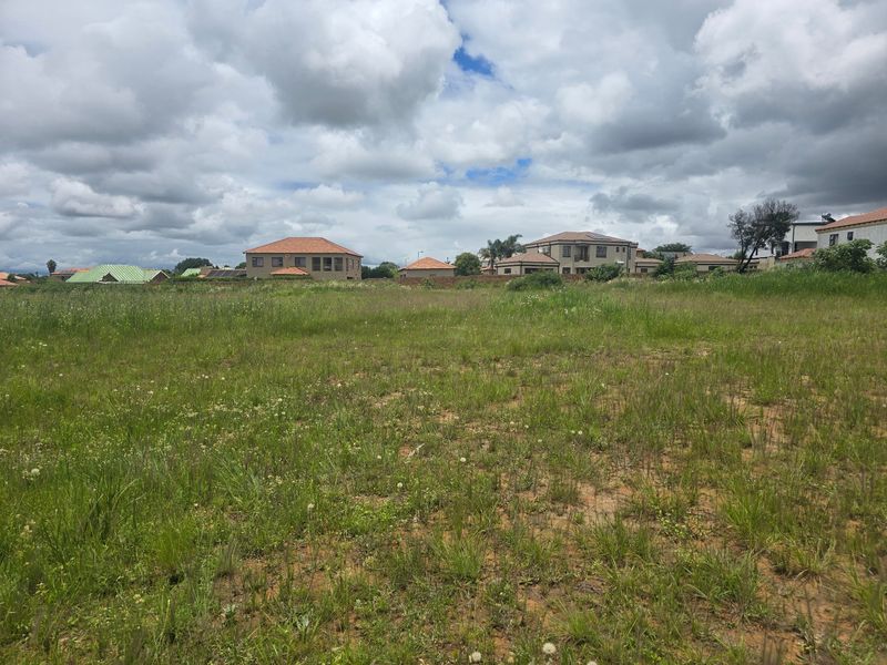 A vacant residential plot of land featuring natural grass coverage and a perimeter wall, situated within an established residential neighborhood.