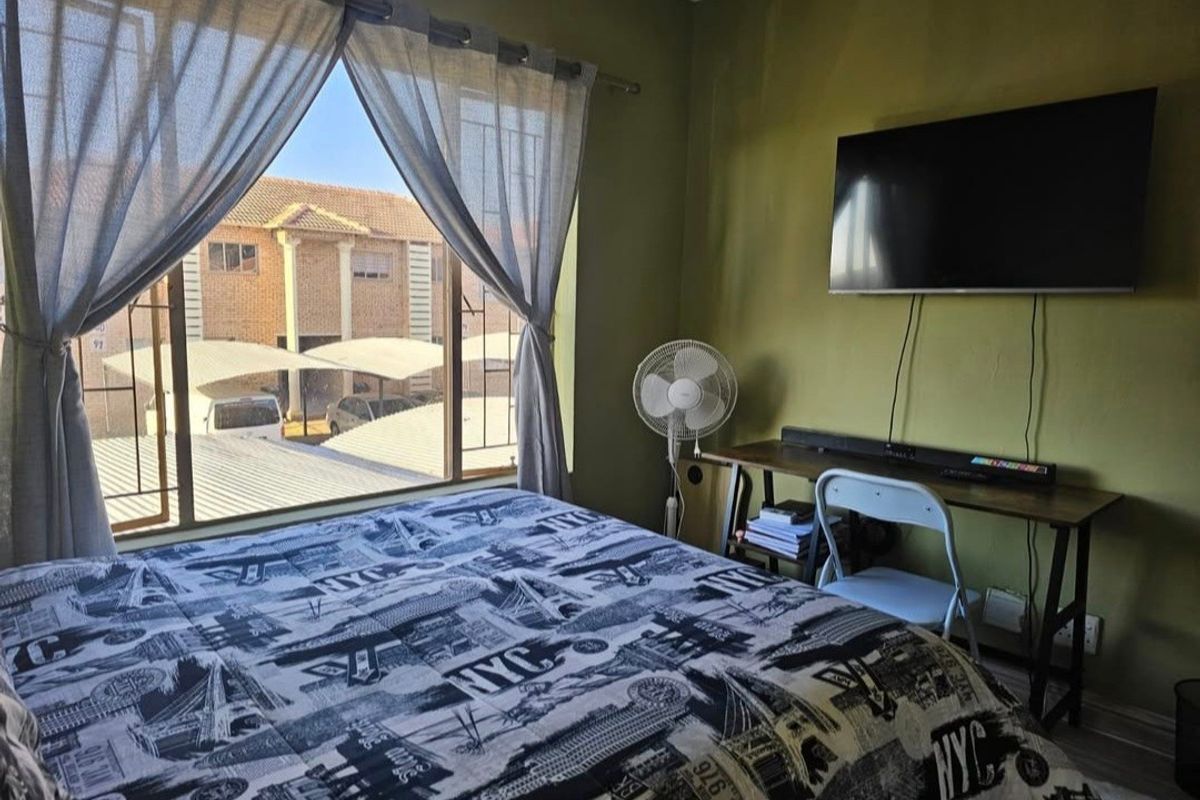 A bedroom featuring a large window with security bars, wall-mounted television, and neutral wall finishes, offering a view of the surrounding complex.