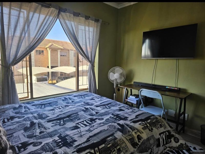 A bedroom featuring a large window with security bars, wall-mounted television, and neutral wall finishes, offering a view of the surrounding complex.