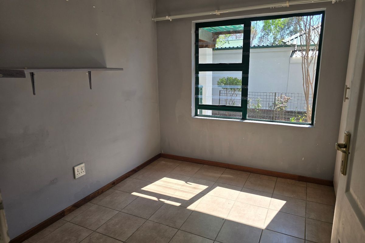 An empty bedroom featuring tiled flooring, a large aluminium window, and a wall-mounted shelf, requiring minor cosmetic updates to the wall finishes.