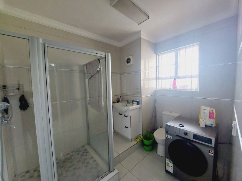 A functional bathroom featuring a glass shower enclosure, vanity basin, toilet, and tiled finishes, with space for laundry appliances.