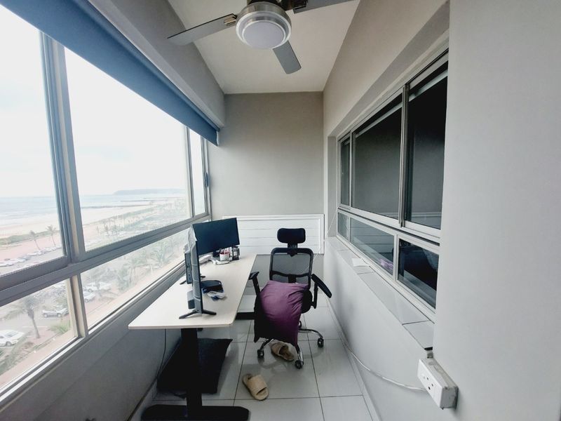 Enclosed balcony space featuring tiled flooring, a ceiling fan, and large windows offering expansive coastal views, currently utilized as a home office.