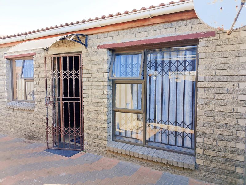 Rear exterior view of the property featuring exposed brickwork, aluminium window frames with security bars, and a paved patio area.