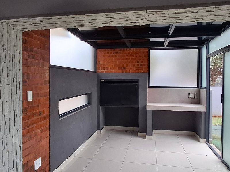 A modern enclosed patio featuring a built-in braai, exposed brick accents, stone cladding, and large sliding glass doors leading to the garden.