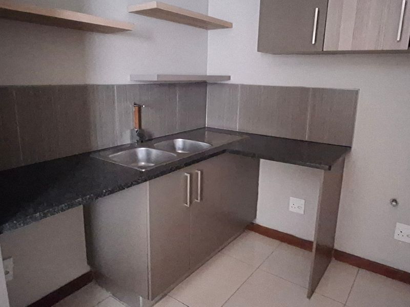 A clean, modern kitchen area featuring built-in cabinetry, granite countertops, a double bowl sink, and floating shelves with neutral tiled flooring.