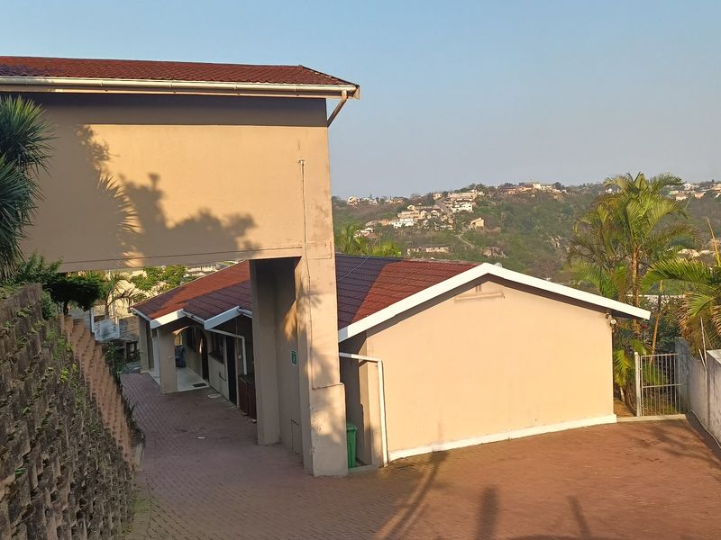 Exterior view of the property featuring a paved driveway, retaining wall, and tiled roof structure with surrounding hillside views.