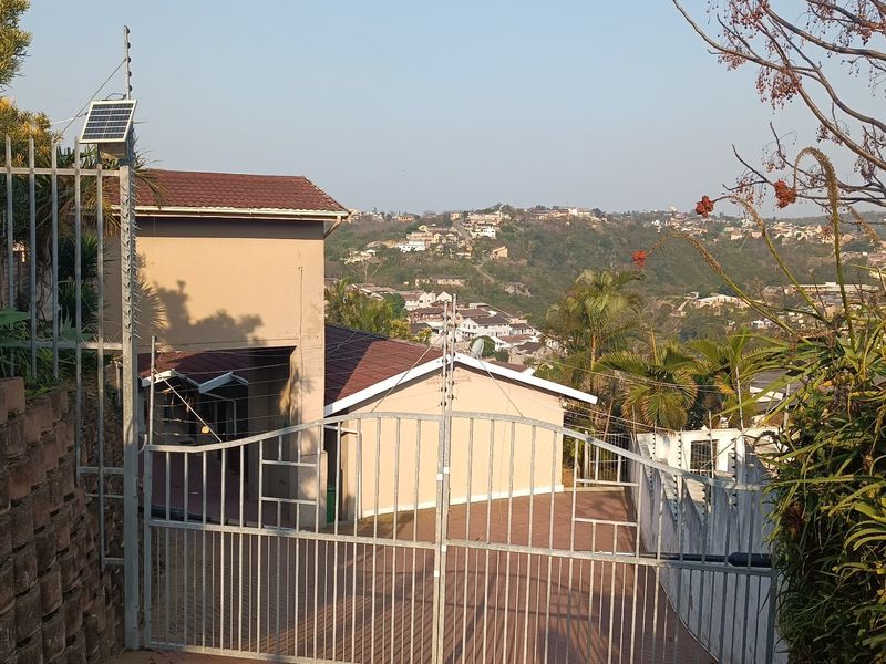 Front entrance view featuring a secure metal gate, electric fencing, solar panel, and a paved driveway leading to the residence with hillside views.