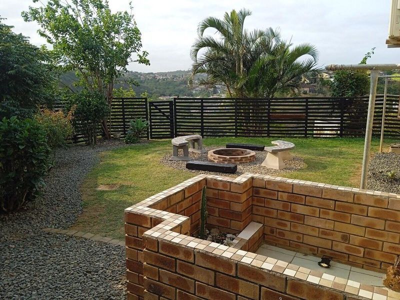 A landscaped backyard featuring a brick fire pit with stone seating, a brick planter box, and a gravel pathway enclosed by a slat fence.