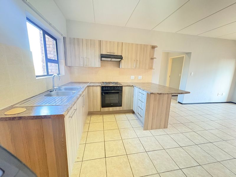 A clean, functional kitchen featuring light wood-look cabinetry, laminate countertops, built-in oven with hob, and tiled flooring in an open-plan layout.
