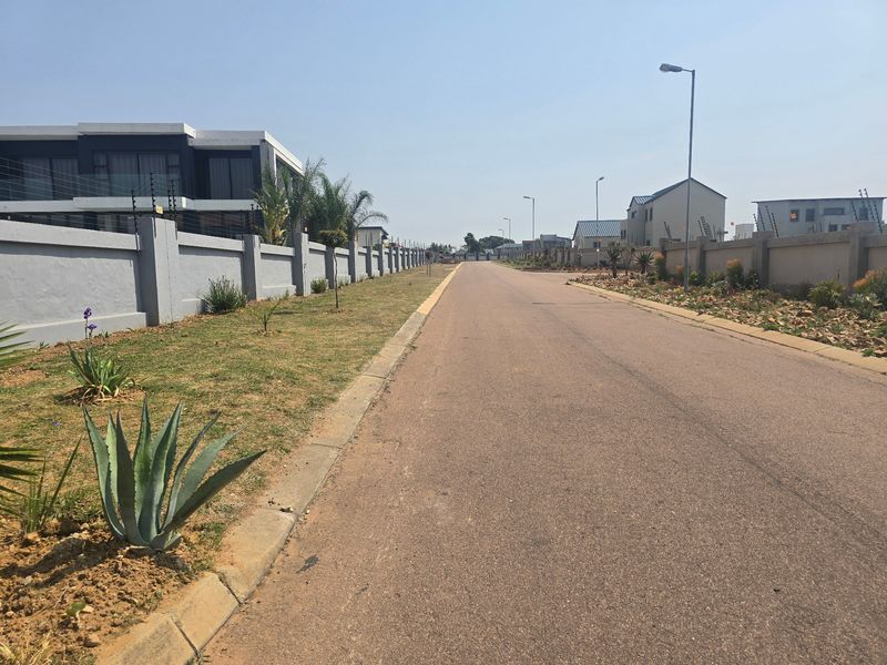 A view of a paved residential street within a secure estate, featuring perimeter walls with electric fencing and established street lighting.