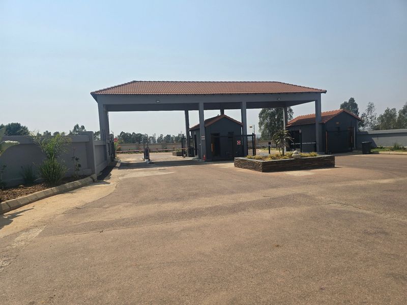 A secure residential estate entrance featuring a gated access point, guardhouse, and paved driveway with a tiled roof structure.