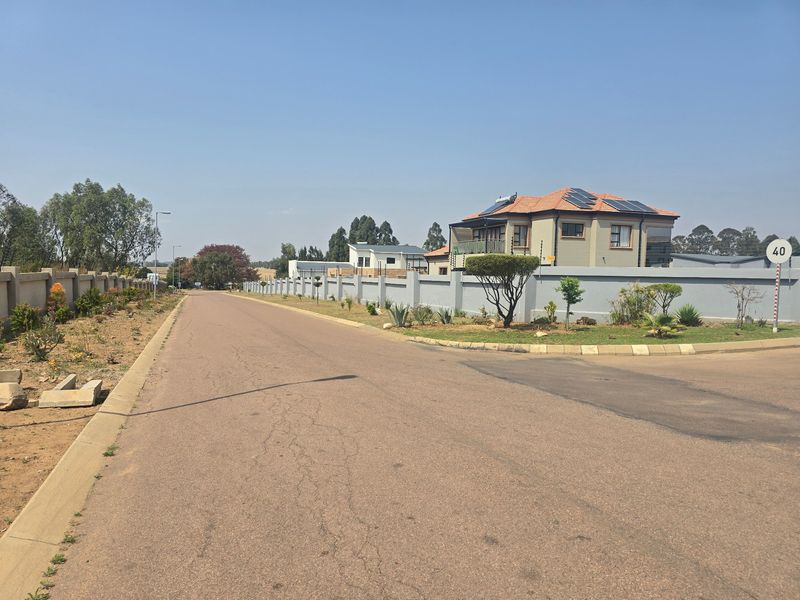 A clear street view of a residential area featuring paved roads, perimeter walls with electric fencing, and houses with solar panel installations.