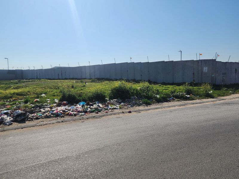 Street view of a vacant residential plot enclosed by a precast concrete boundary wall with electric fencing, currently requiring significant site clearing.