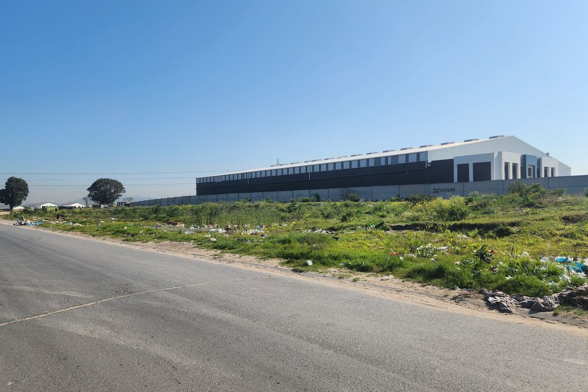 Street view of a vacant residential plot adjacent to an industrial warehouse facility with an asphalt road frontage.