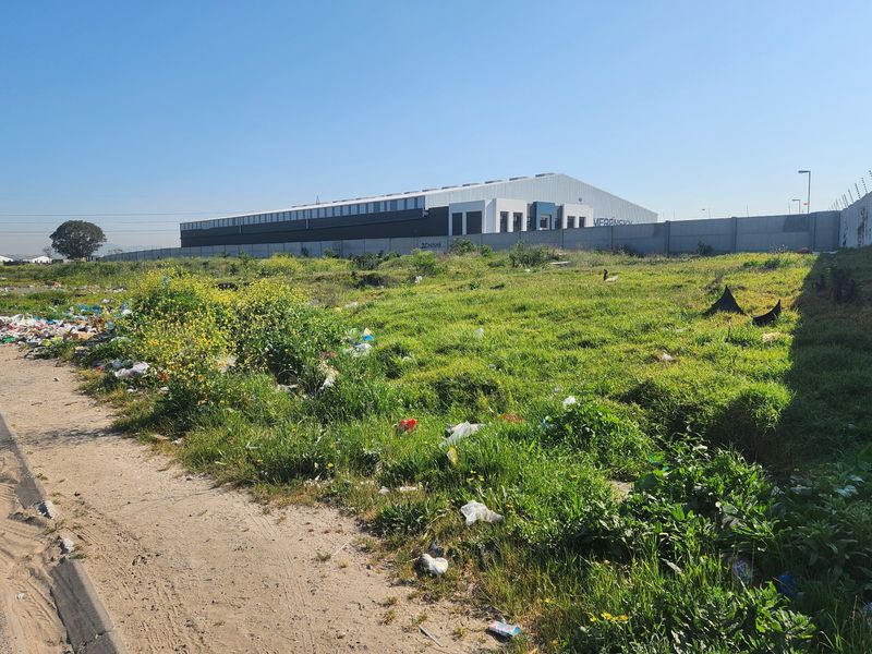 Vacant residential land plot featuring overgrown grass and perimeter walling, situated adjacent to a commercial warehouse facility.