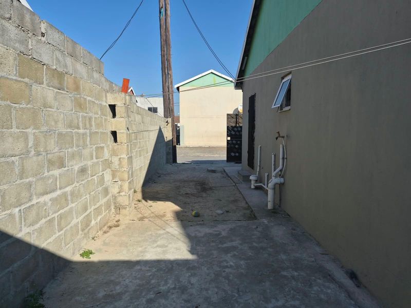 A narrow side or rear exterior passage featuring a concrete block boundary wall, paved ground, and exposed external plumbing fixtures.