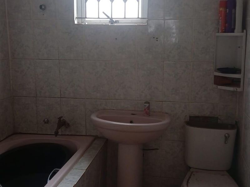 A dated bathroom featuring a pedestal basin, built-in bathtub, and toilet with tiled walls and a security-gated window.