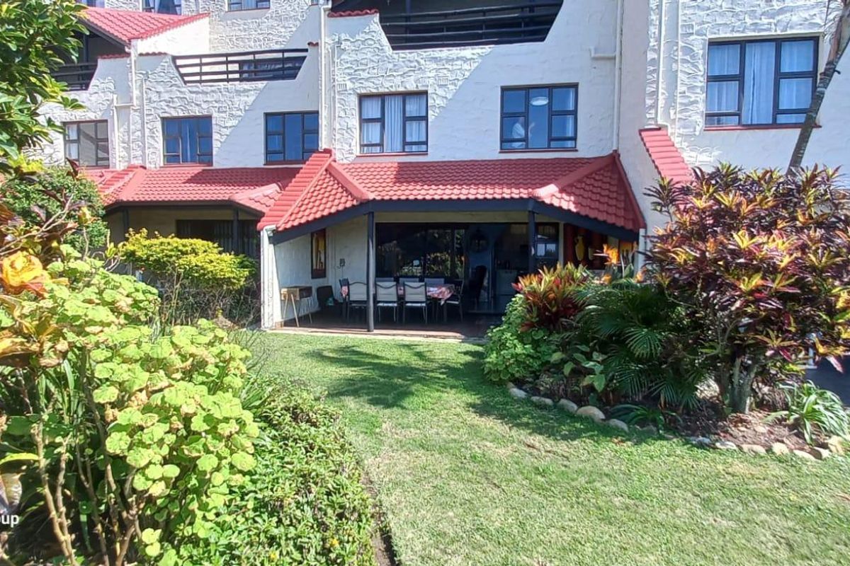 Rear view of the apartment complex featuring a covered patio area, tiled roofing, and a well-maintained private garden space.