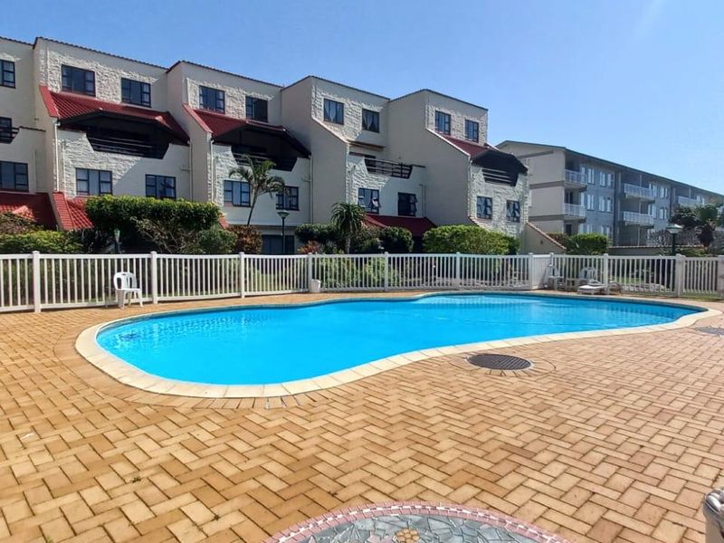 A communal swimming pool area featuring paved surrounds and a white picket fence, set against the backdrop of a multi-story apartment complex.