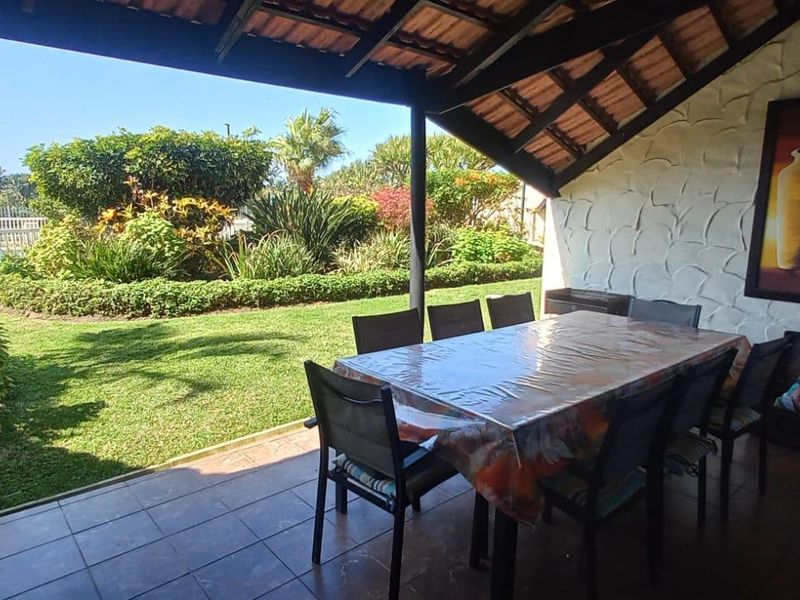 A covered patio area featuring tiled flooring and exposed timber rafters, overlooking a lush garden and pool area.