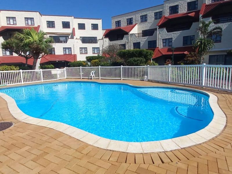 A well-maintained communal swimming pool area featuring paved surrounds and a safety fence, set against the backdrop of a multi-story apartment complex.