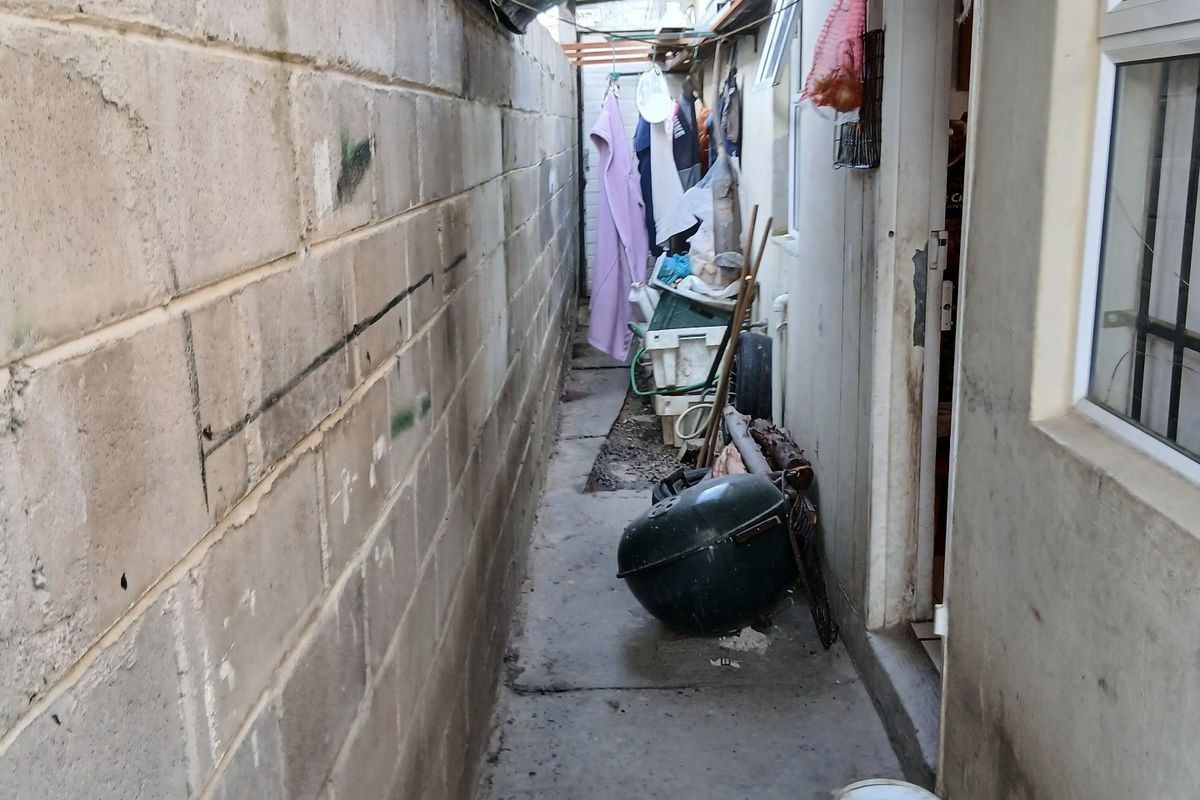 A narrow side passage featuring a concrete block boundary wall and concrete pathway, currently used as a utility and storage area.