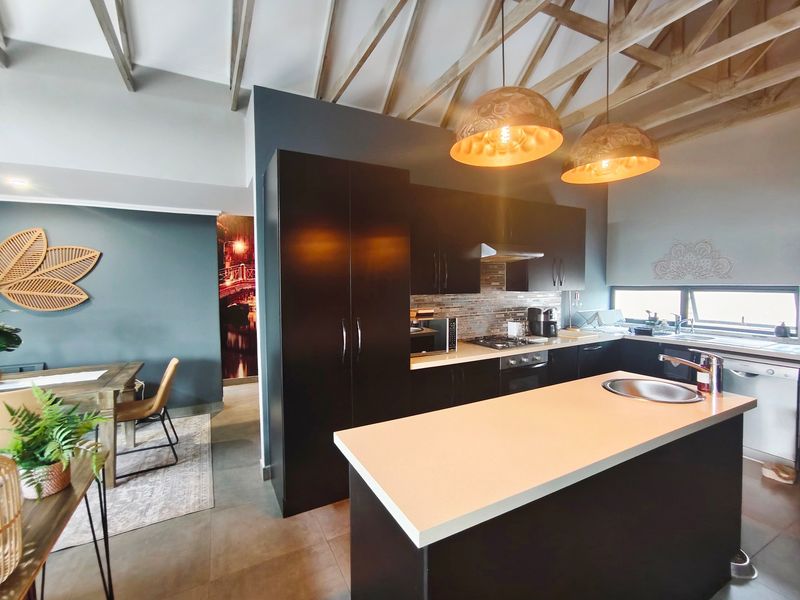 Modern open-plan kitchen and dining area featuring exposed timber rafters, dark cabinetry, stone countertops, and a functional kitchen island with a prep sink.