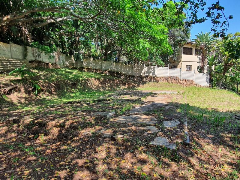 A sloped residential plot featuring mature trees, a precast concrete boundary wall, and a stone retaining wall, offering potential for landscaping or development.