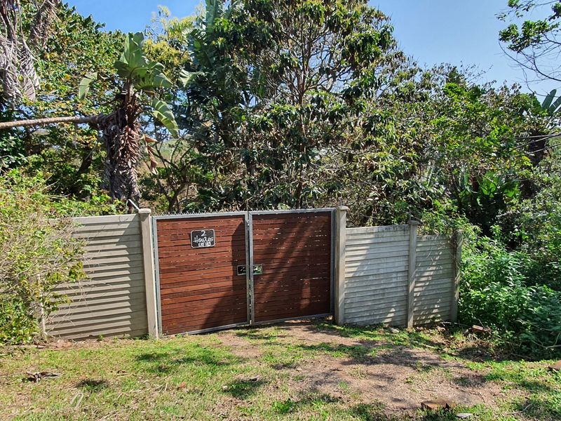 A view of the property entrance featuring a timber gate set within a precast concrete boundary wall, surrounded by established trees and vegetation.