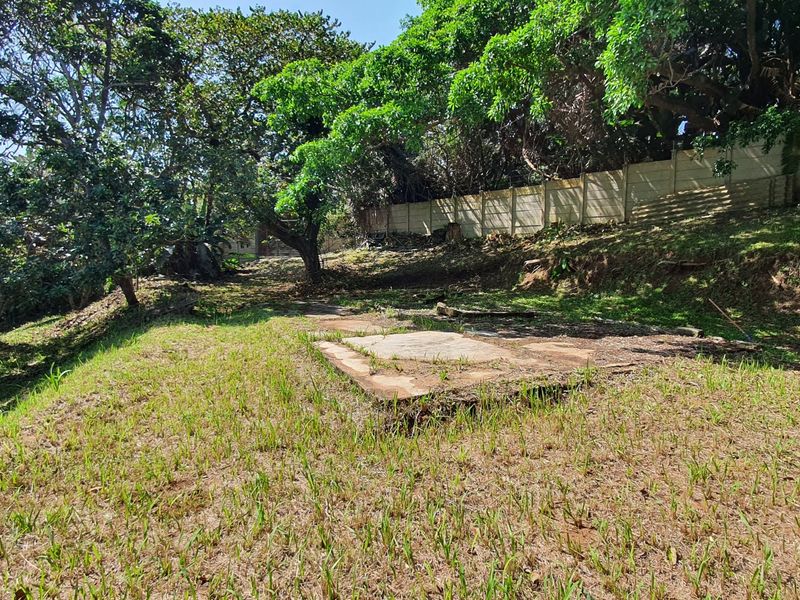 A sloped residential plot featuring mature trees, a concrete slab foundation, and a perimeter retaining wall.