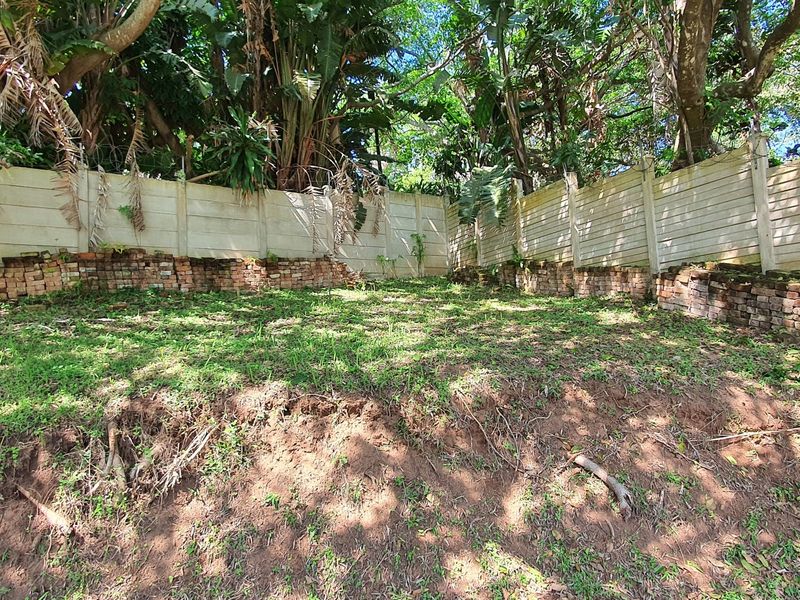 A vacant residential plot featuring a precast concrete boundary wall, mature trees, and a low brick retaining wall along the perimeter.