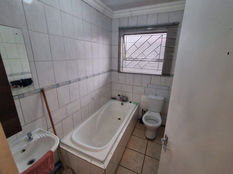A functional bathroom featuring a built-in bathtub, pedestal basin, and toilet, finished with ceramic wall and floor tiles and a window with security bars.