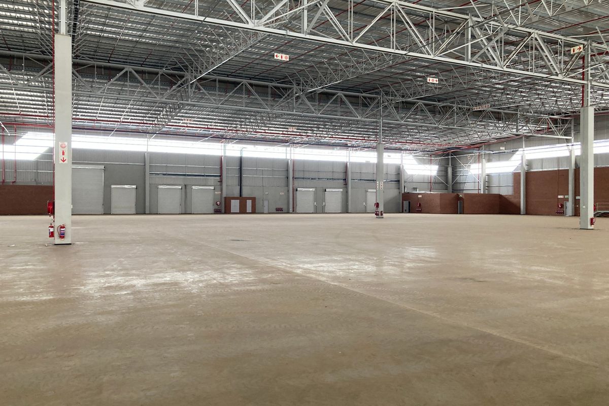 Spacious industrial warehouse featuring high ceilings with exposed steel trusses, concrete flooring, multiple roller shutter doors, and an integrated fire suppression system.