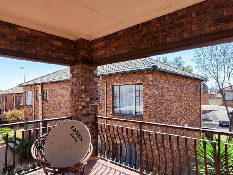 A covered balcony with brick walls and metal railings, overlooking the apartment complex grounds.