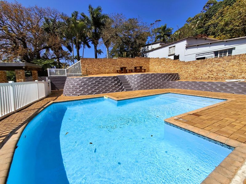 Outdoor pool area featuring a paved deck, tiered brick retaining walls, and white perimeter fencing, set against a landscaped backdrop.