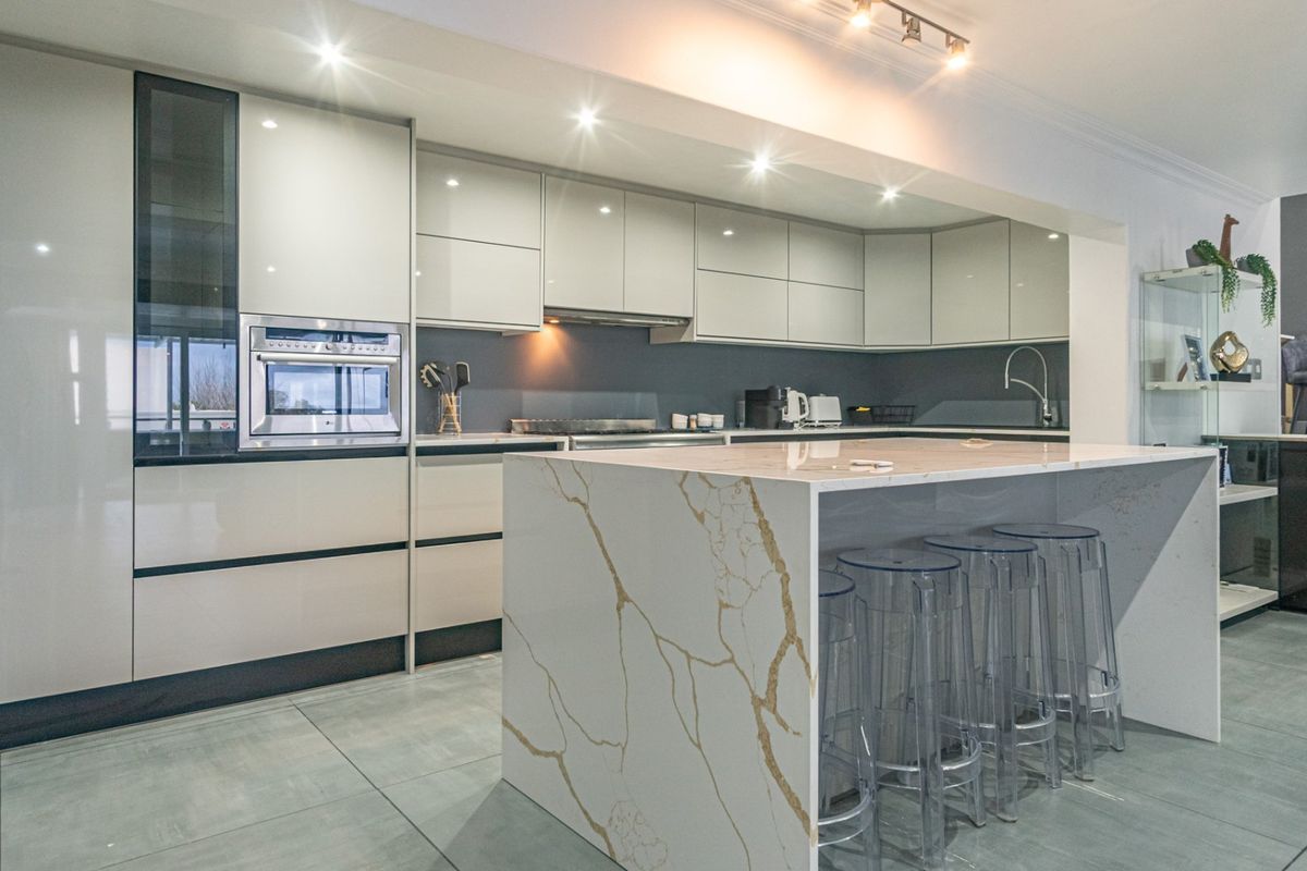 Modern kitchen featuring high-gloss cabinetry, a large stone-topped island, built-in oven, and sleek tiled flooring with recessed lighting.