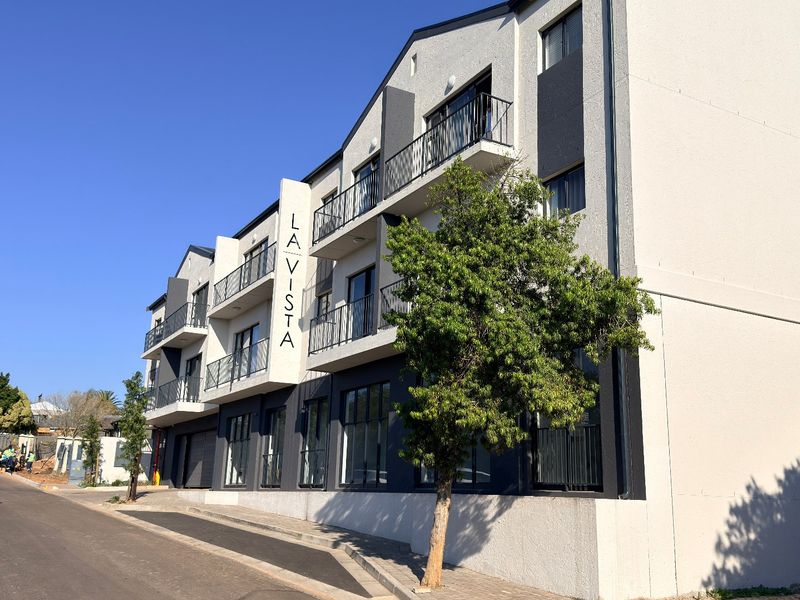Modern multi-story apartment building facade featuring balconies with metal railings and large windows, situated along a paved street.