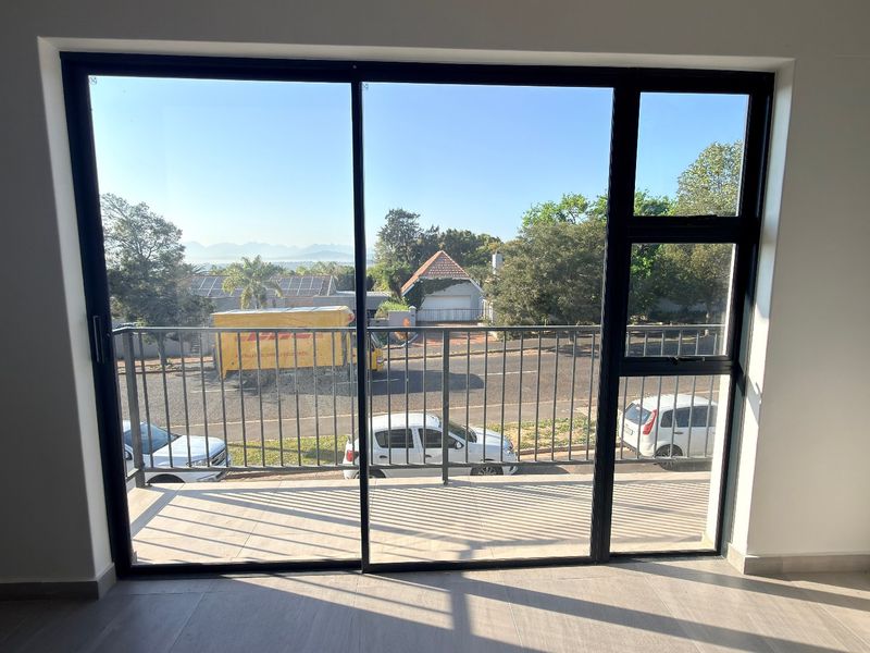 A bright, modern balcony featuring a sleek metal balustrade and large sliding glass doors, offering an elevated view of the surrounding street and neighborhood.