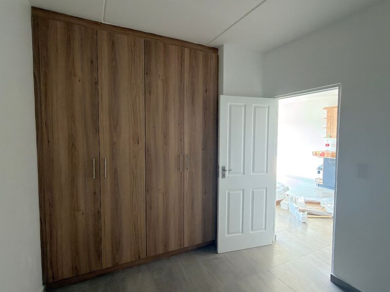 A clean, empty bedroom featuring modern wood-finish built-in cupboards and neutral tiled flooring, with a view into the adjacent living area.