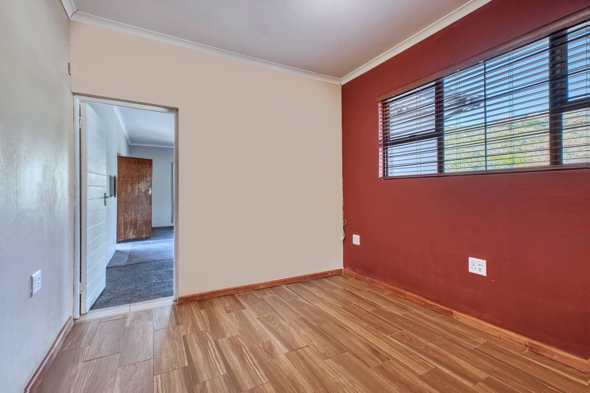 An empty bedroom featuring wood-look laminate flooring, a feature wall with venetian blinds, and direct access to an adjacent hallway.
