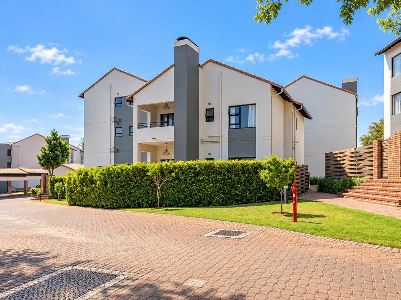 A well-maintained townhouse exterior featuring a paved driveway, brick retaining walls, and private balconies set within a landscaped residential complex.